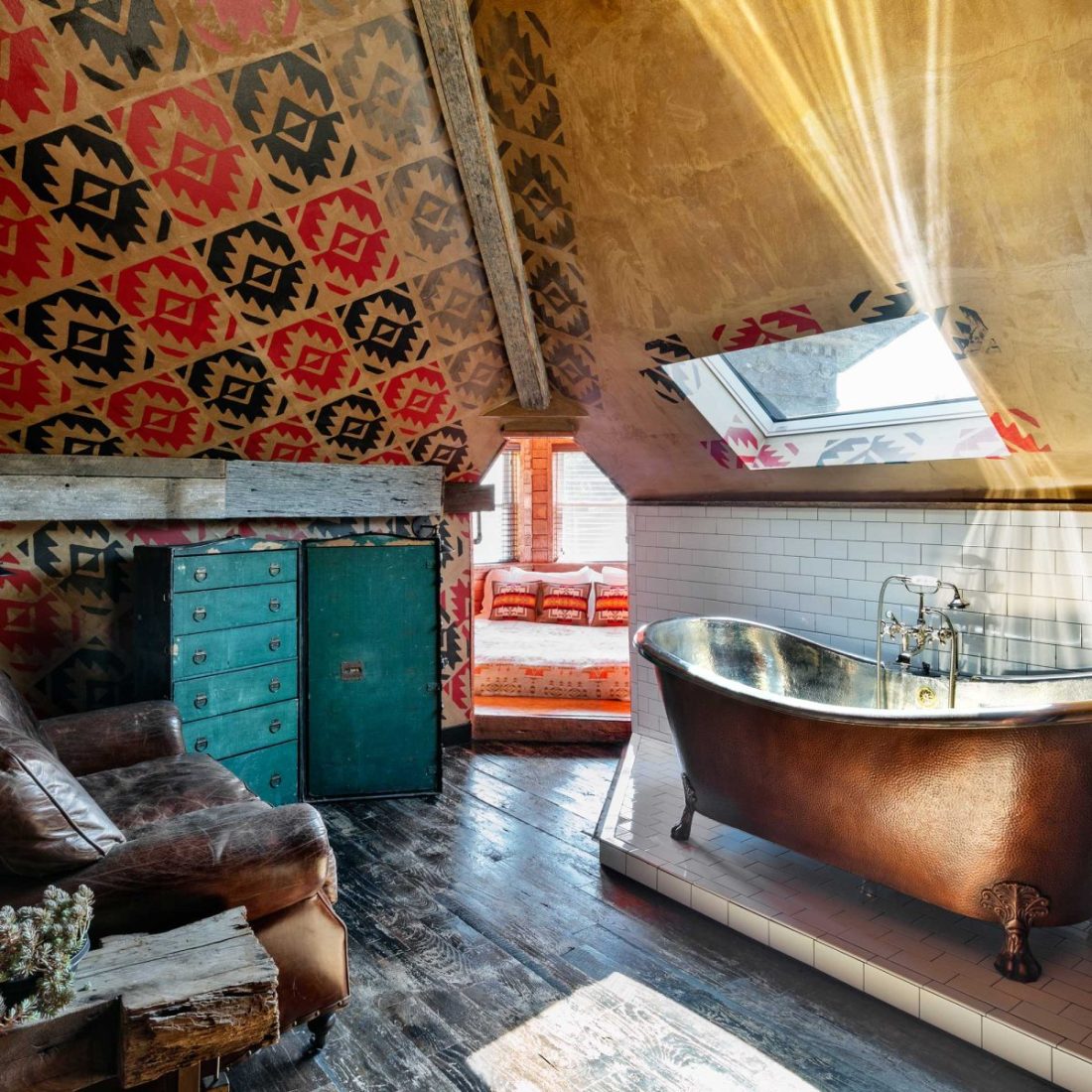 Entering The Tower room at Urban Cowboy Nashville. A comfortable couch is placed, a set of drawers beside. A small ingress leads to a windowed tower area for the bed. Across is a raised tile area holding a copper claw foot tub and stool.