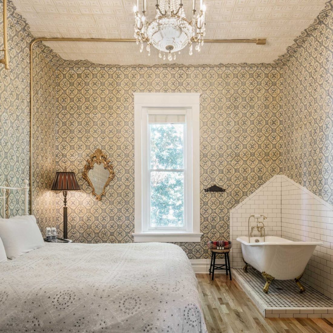 The Victorian Room inside Urban Cowboy Nashville, lit with a warm light of the chandelier. A king sized bed is placed in the middle, just across from a tiled area holding a claw foot bathtub.