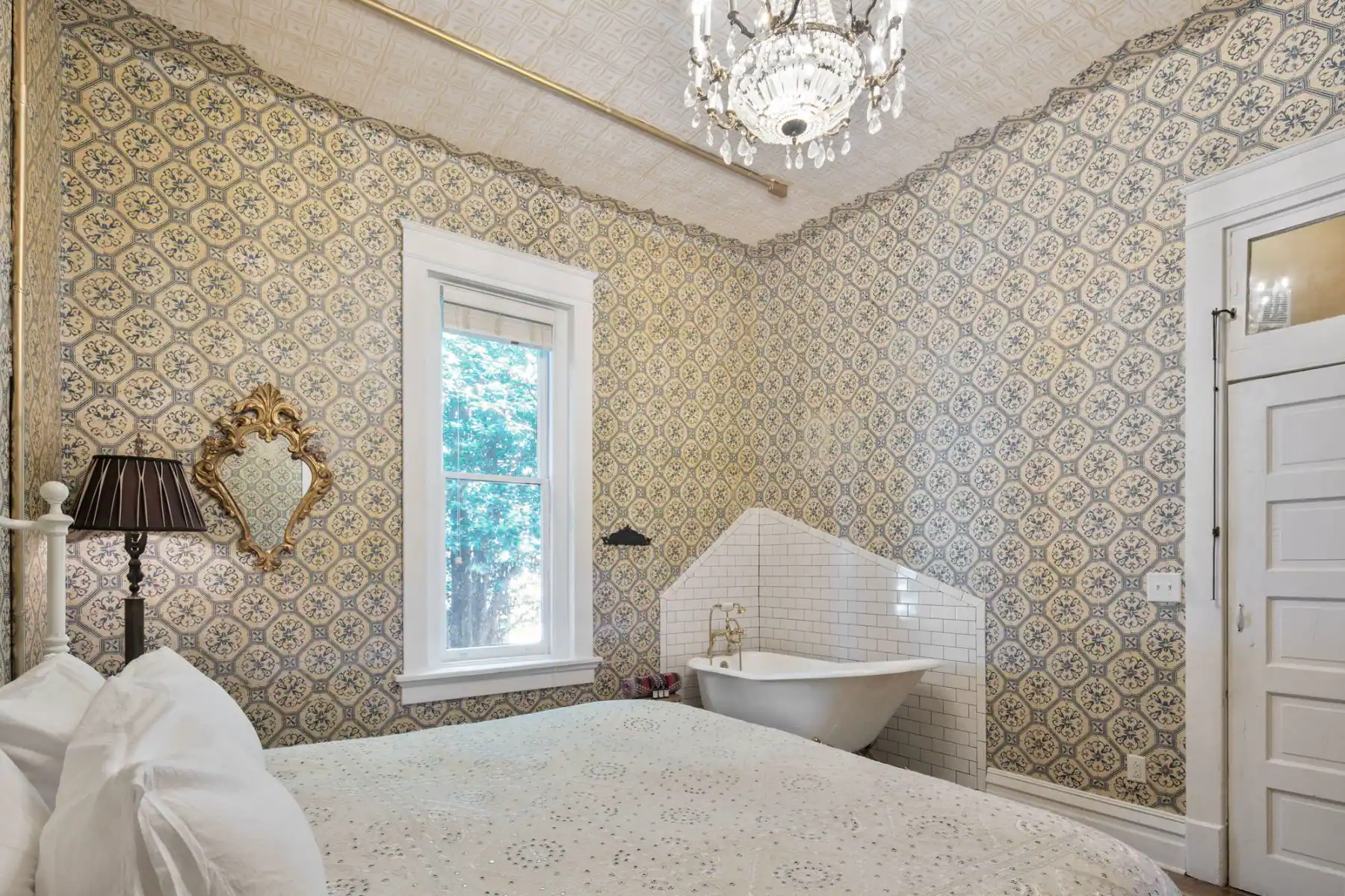 View from beside the king sized bed inside The Victorian Room of Urban Cowboy Nashville. Across from you is a tiled corner, holding a claw foot bathtub. A Victorian floor lamp is near the window, alongside a matching wall mirror.