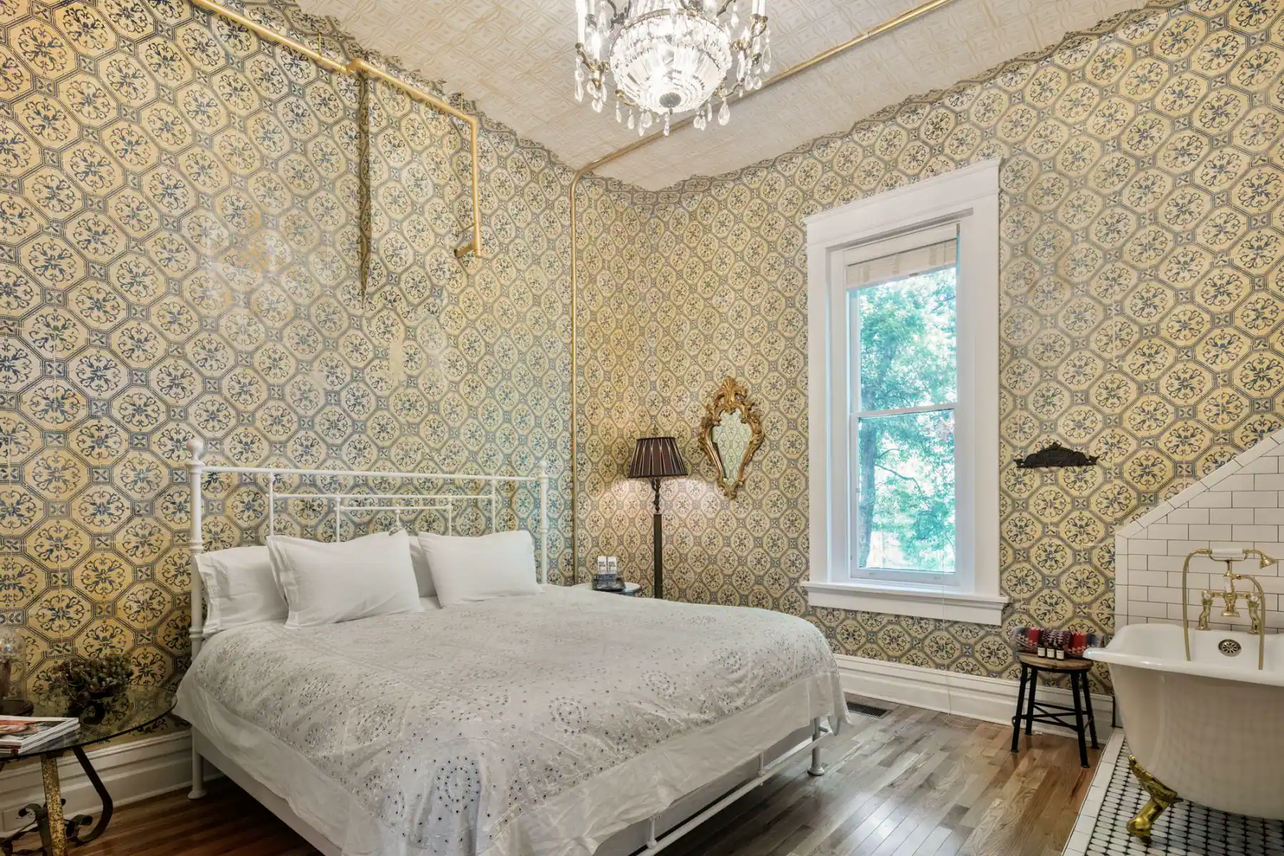 The Victorian Room inside Urban Cowboy Nashville. A king sized bed is placed in the middle, just across from a tiled area holding a claw foot bathtub. The walls are tiled with detail and a chandelier lamp gives a warm light