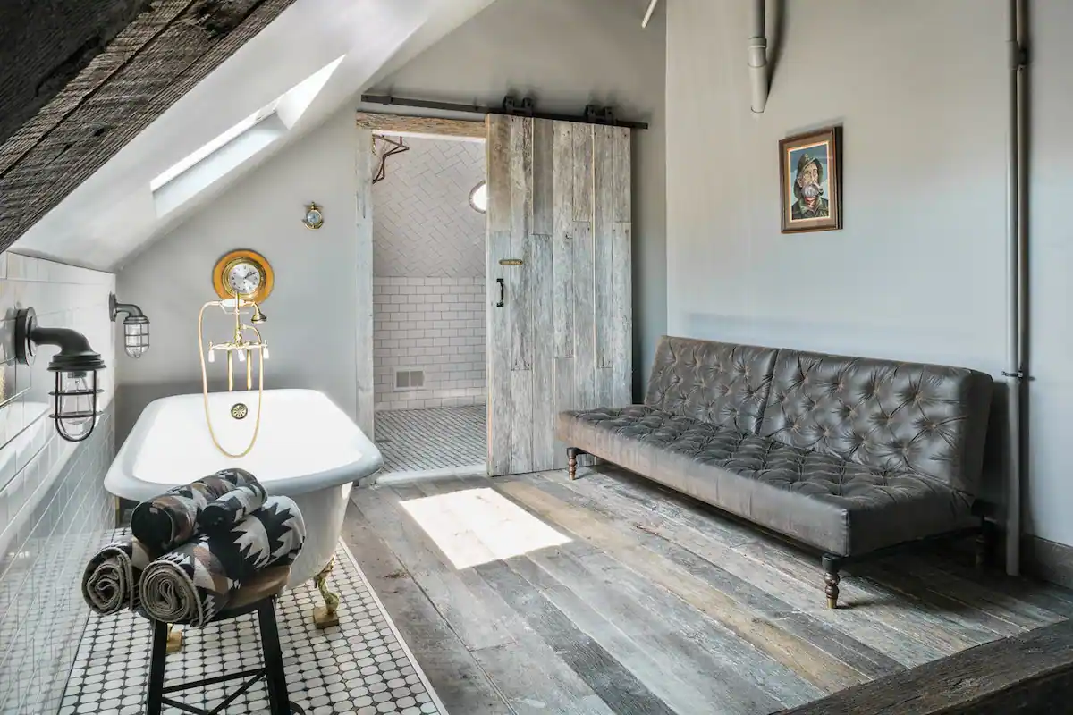 Looking into the bathing area and shower room inside The Captain Suite of Urban Cowboy. The king sized bed is tucked into the hull, with a porthole window. A claw foot bathtub is nearby, beneath a skylight.