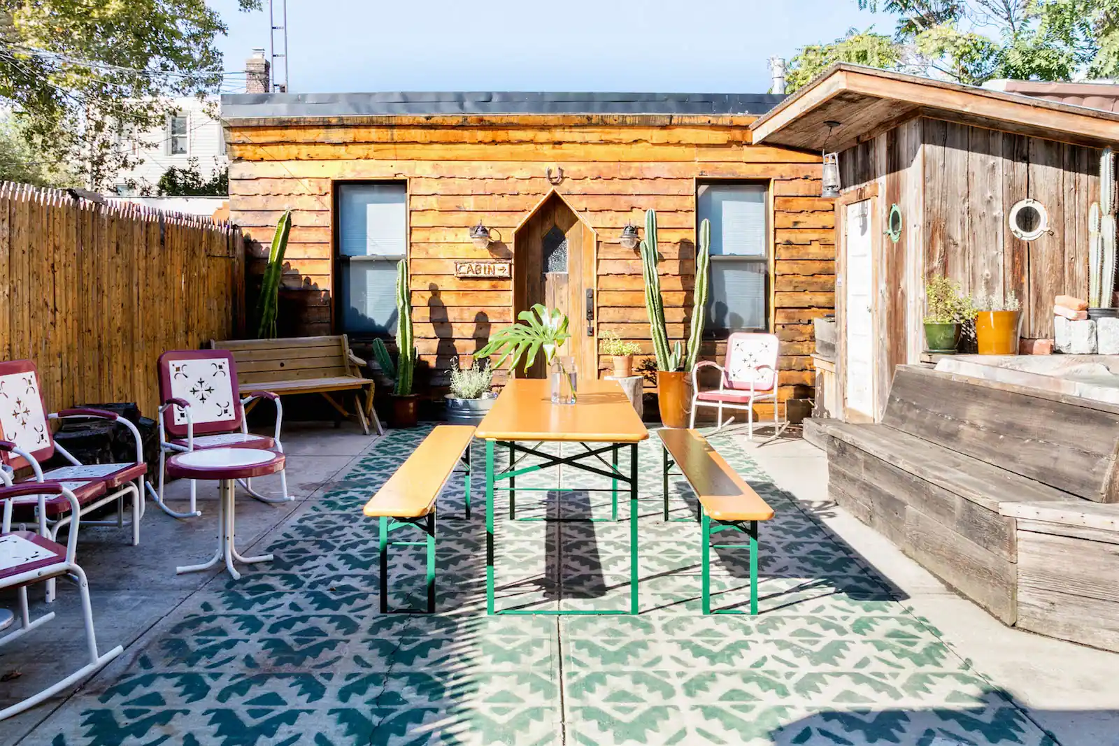 Fenced in back patio area of Urban Cowboy Brooklyn. Green tile cuts a pattern on the ground, with chairs scattered around the table and bench.